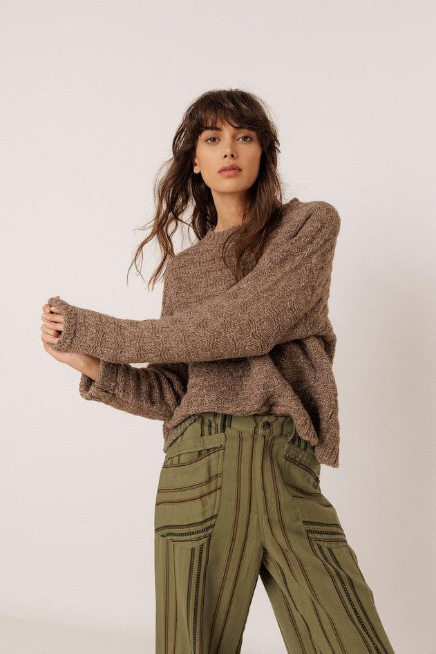A woman modeling a brown chunky knit sweater and olive green striped trousers against a plain light background.