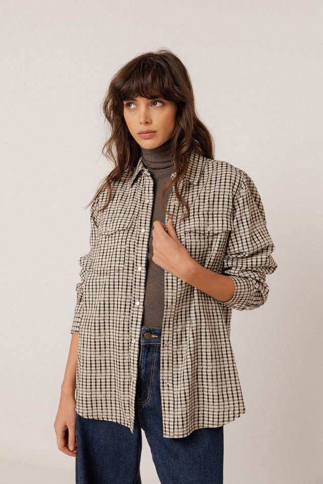 A woman modeling a beige and black checkered button-up shirt over a dark gray turtleneck sweater and blue jeans, photographed against a plain light gray studio background.