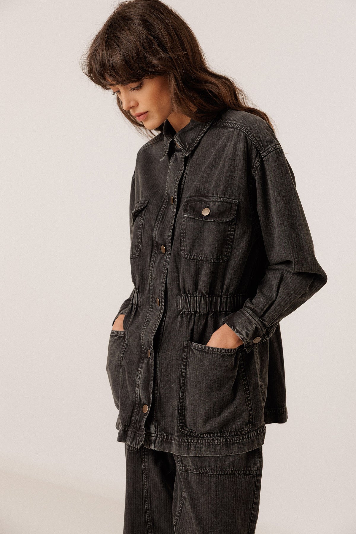 A woman wearing a dark charcoal gray corduroy utility jacket with front pockets and a drawstring waist, paired with matching dark corduroy pants, photographed against a plain light background.