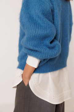 A close-up of a person wearing a fuzzy blue knit sweater layered over a white blouse with a scalloped hem, paired with gray trousers, against a plain light background.