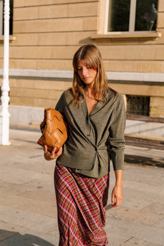 A woman walking outdoors on a sunny day, wearing a dark olive green button-up shirt and a red and green plaid skirt, while holding a brown woven leather clutch bag.