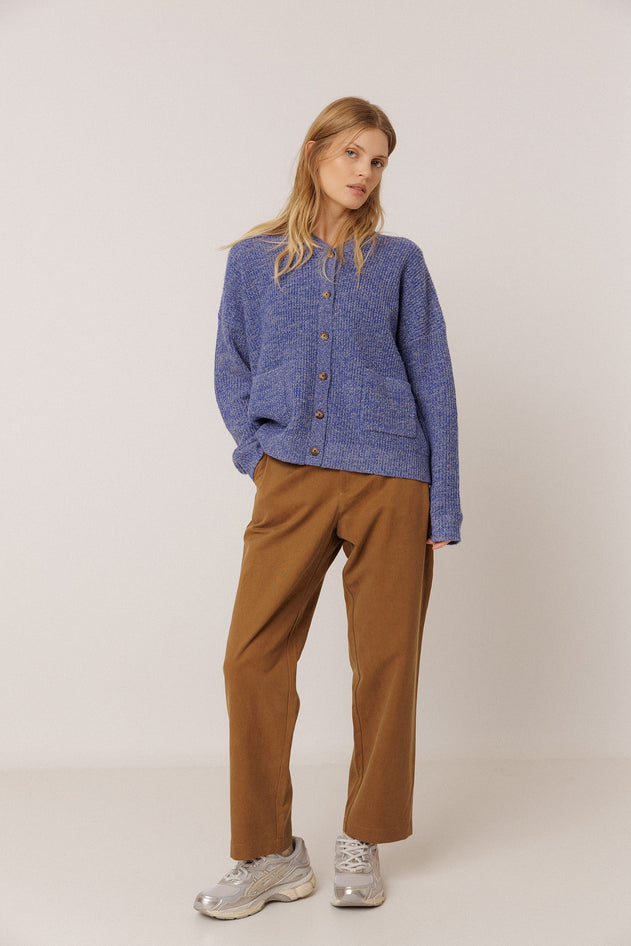 A woman modeling a blue textured button-up cardigan sweater, brown wide-leg trousers, and silver sneakers against a plain light gray studio background.