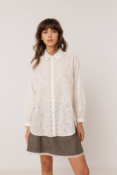 A woman modeling a white eyelet long-sleeve button-up shirt with a pointed collar, paired with a brown textured mini skirt, against a plain light gray studio background.