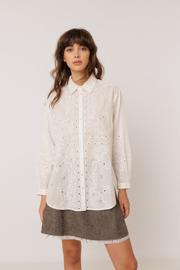 A woman modeling a white eyelet long-sleeve button-up shirt with a pointed collar, paired with a brown textured mini skirt, against a plain light gray studio background.