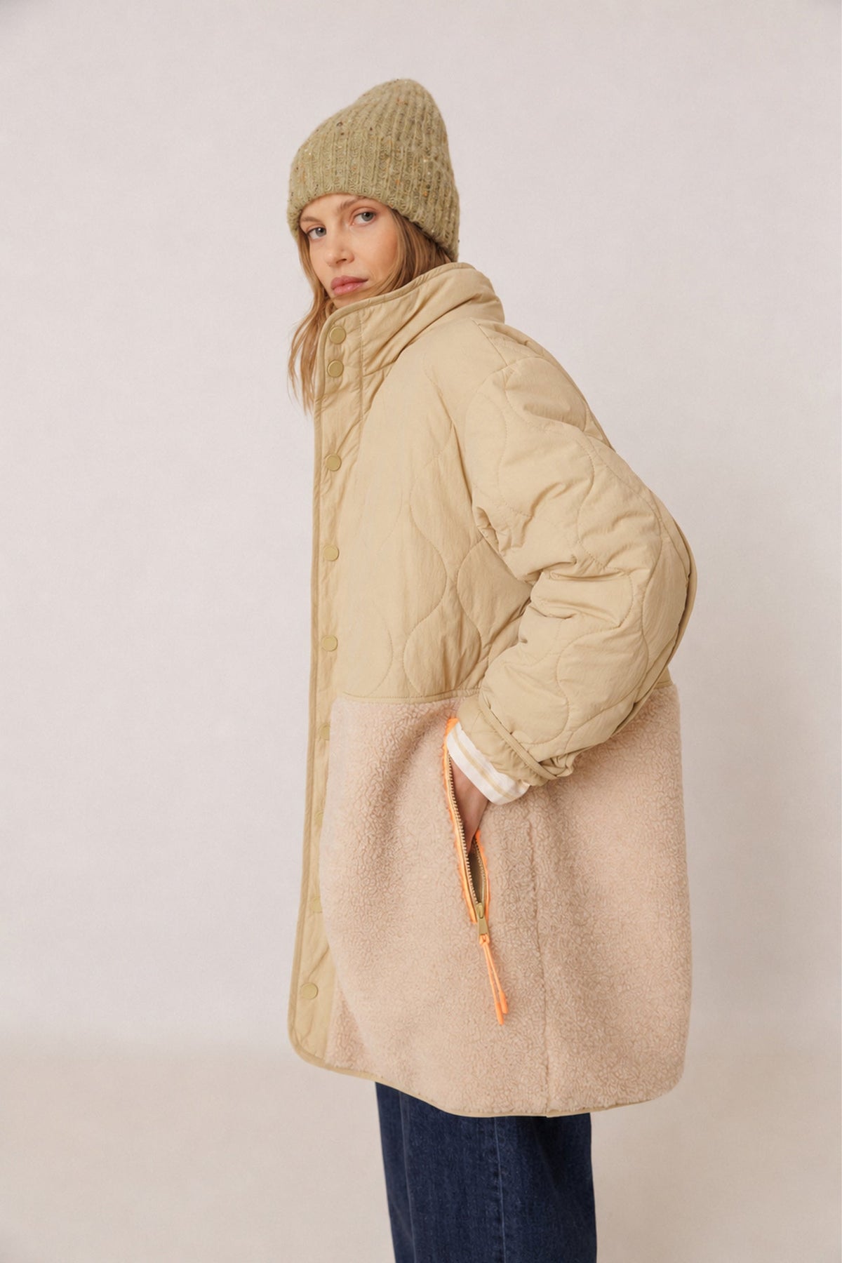 A woman wearing a beige quilted puffer jacket with a soft beige sherpa lower panel, a beige knit beanie, and blue jeans, photographed against a plain light beige studio background.