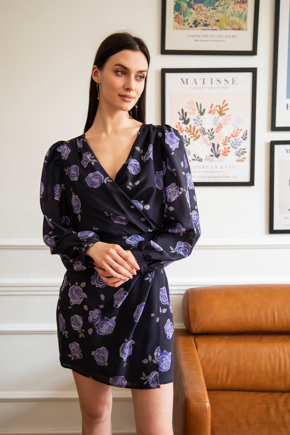 A woman standing in a room with framed pictures on the wall behind her, wearing a navy blue long sleeve mini dress with a rose pattern and a V-neckline. The dress has a closed envelope cut and is fastened on the side with a covered zipper.