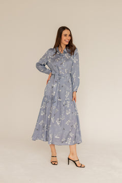 A woman standing in a studio setting, wearing a blue long sleeve maxi dress with a floral pattern, buttoned at the waist, with a belt, and a small collar.